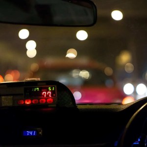 Nighttime view from inside a car, focusing on the illuminated taxi meter showing a fare of 77. Blurred city lights and traffic through the windshield echo the birth of capitalism, with each light representing commerce in motion.