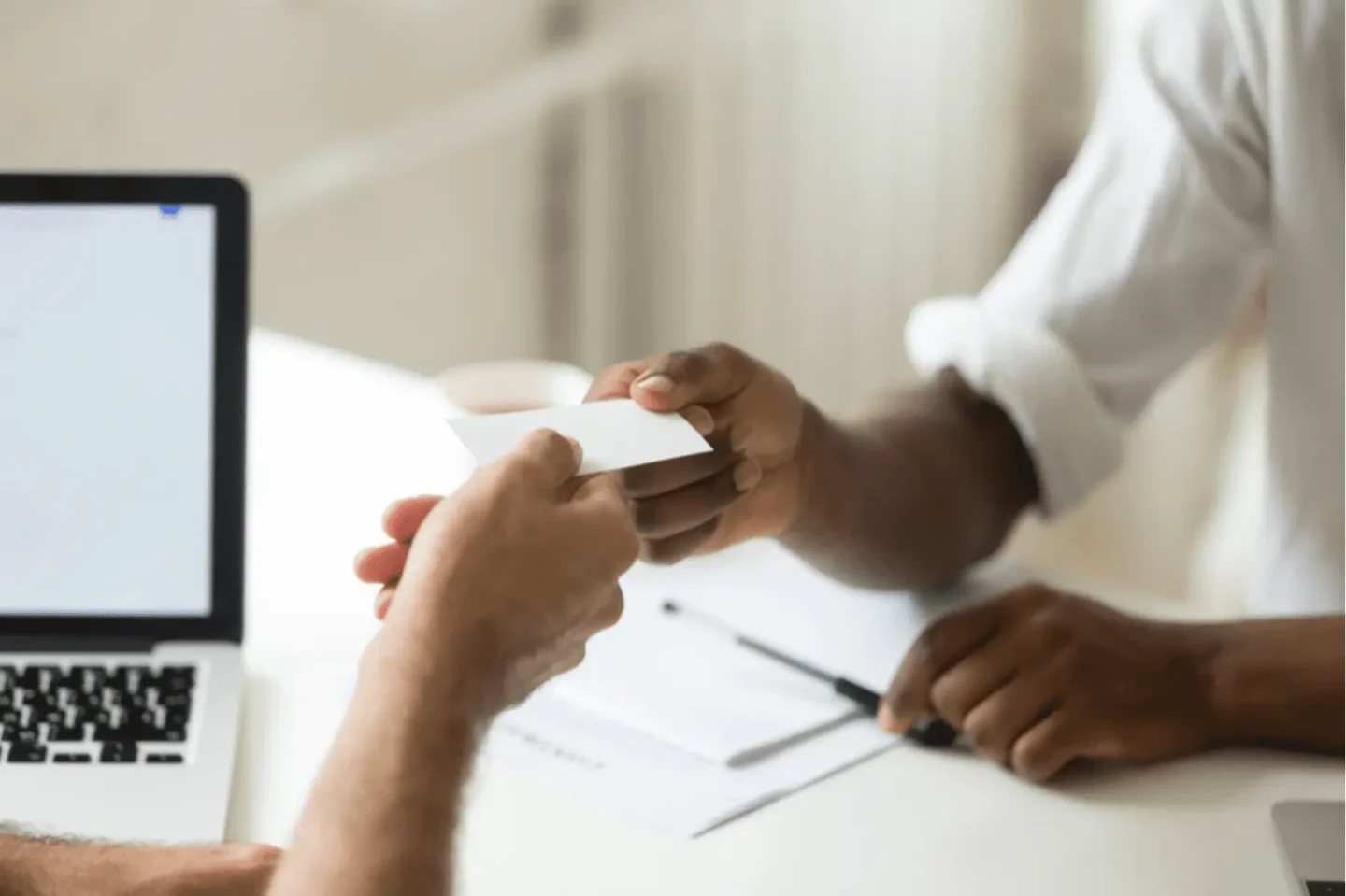 Close-up of two people exchanging a blank business card at a desk, with a laptop and documents visible in the background. One person is handing the card, while the other is reaching to receive it.