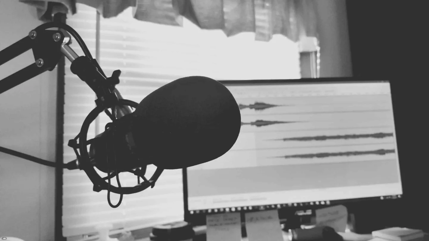 A close-up of a microphone on a boom arm in front of a computer monitor displaying podcast audio waveforms, with sunlight streaming through window blinds in the background.
