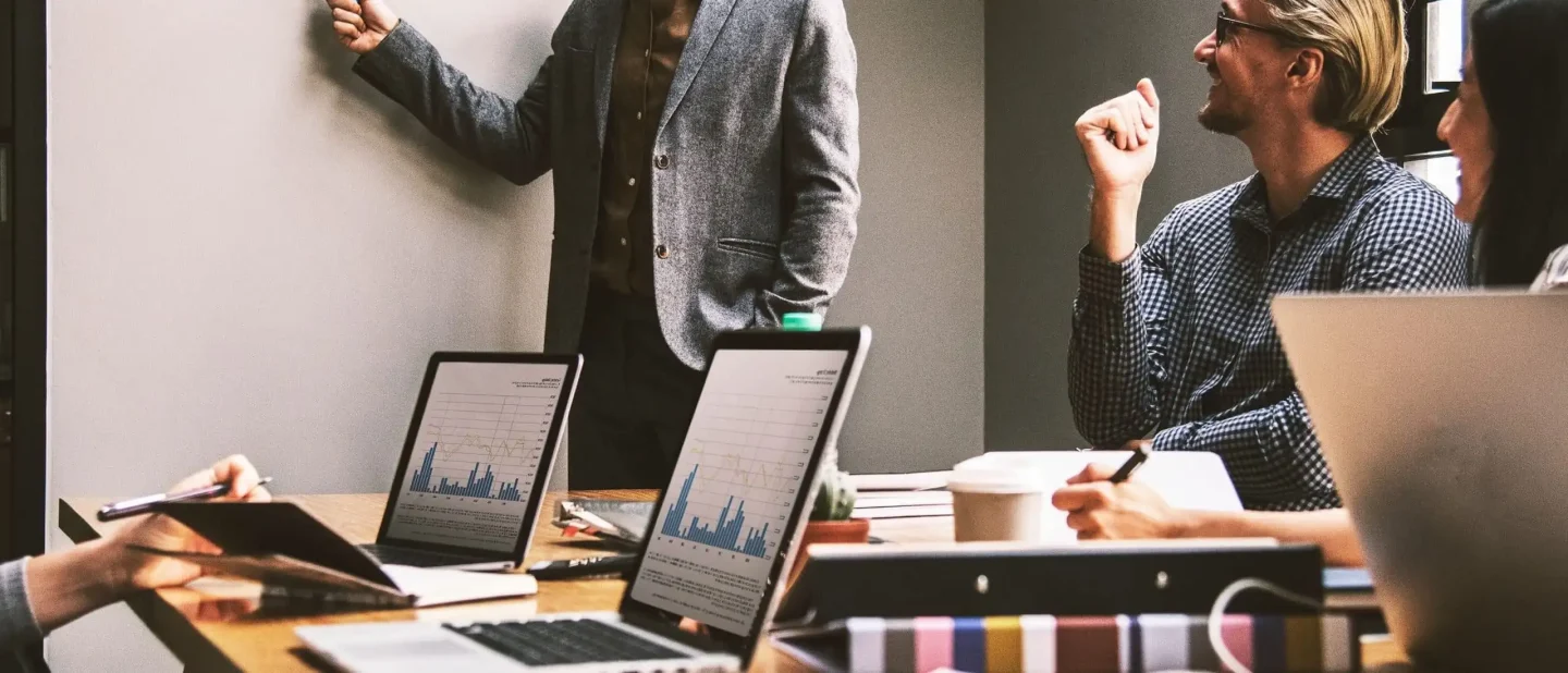 A group of people in a meeting room with laptops showing graphs, one person standing and pointing at a whiteboard explaining strategies to boost my business, while others sit, listen, take notes, and discuss ideas.