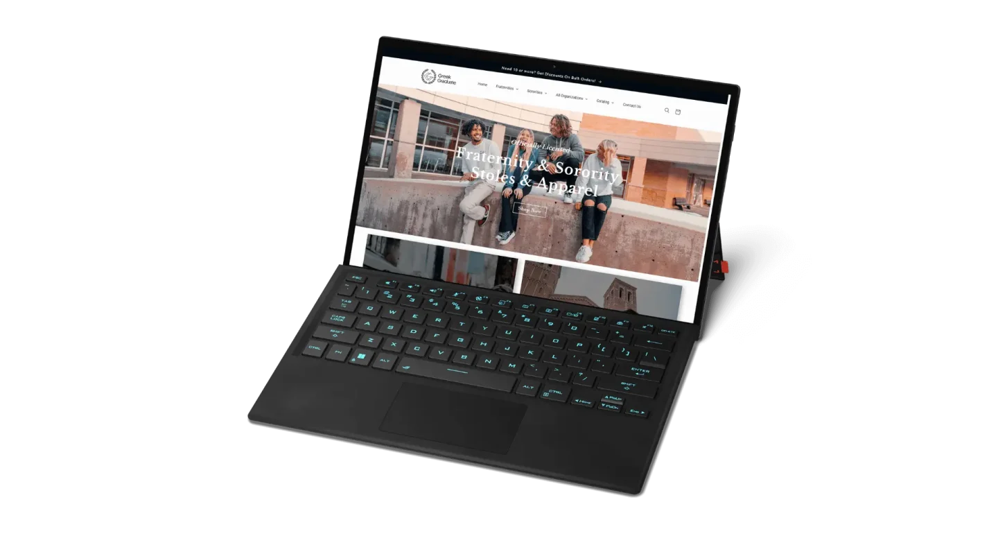 A black laptop with a backlit keyboard displays a website featuring three people sitting on steps and the text Greek Graduate Fraternity & Sorority Stoles & Paper on the screen.
