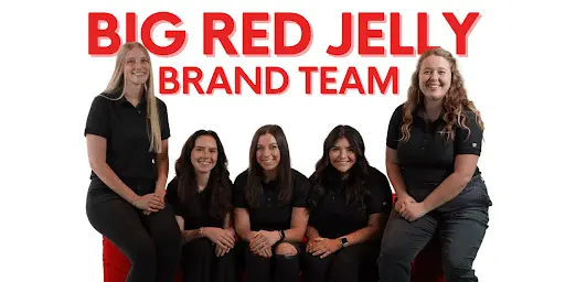 Five women dressed in black sit and stand before a white background with bold red text reading “BIG RED JELLY BRAND TEAM.” Smiling and posing together, they embody Breaking Barriers in Branding.