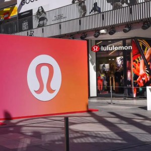 A large sign with the Lululemon logo stands in front of a Lululemon store, with people walking outside and other store signs visible in the background.