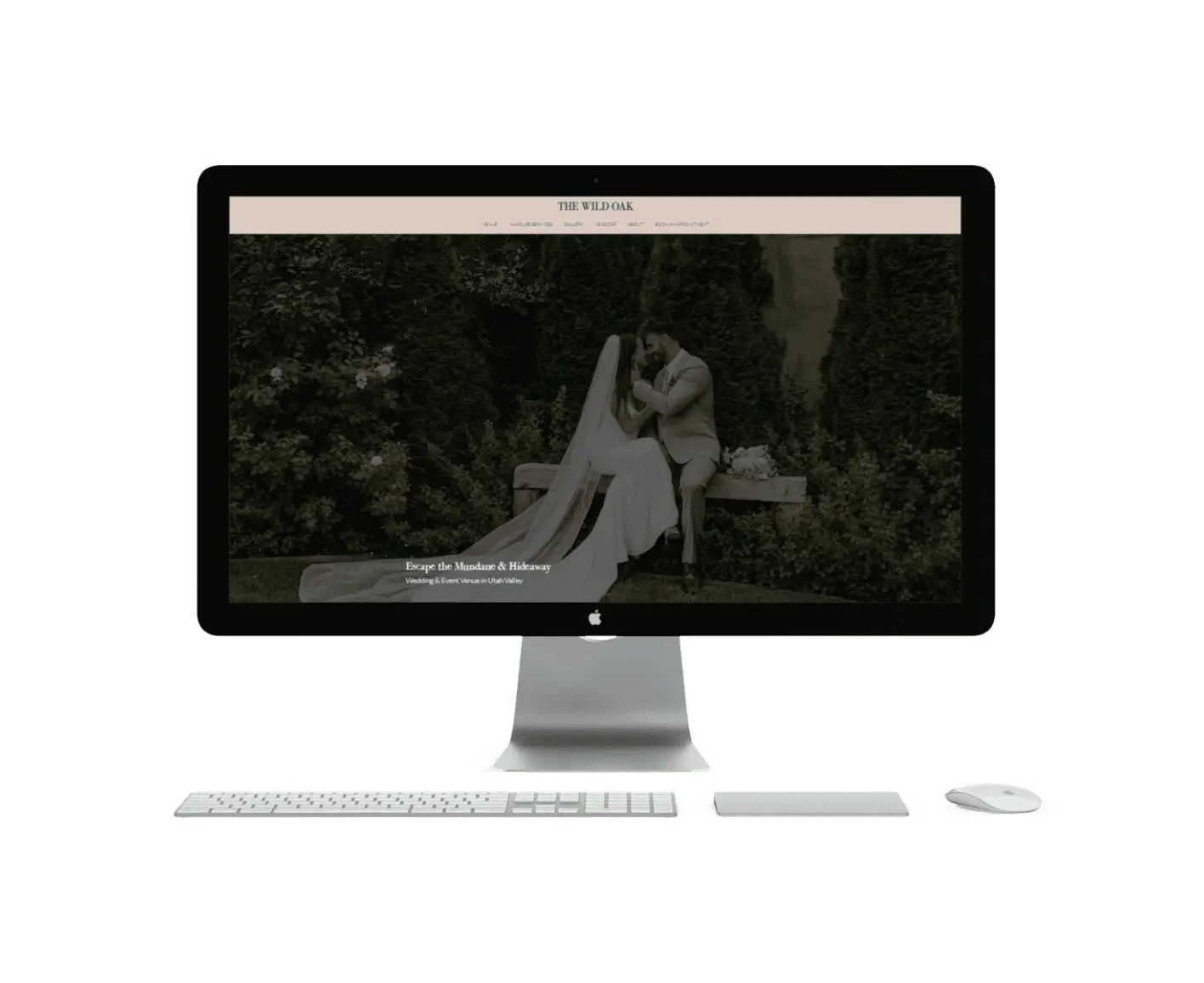 A desktop computer displays a wedding website homepage for Wild Oak Venue, featuring a black-and-white photo of a bride and groom sitting on a bench and kissing, with a keyboard and mouse in front.