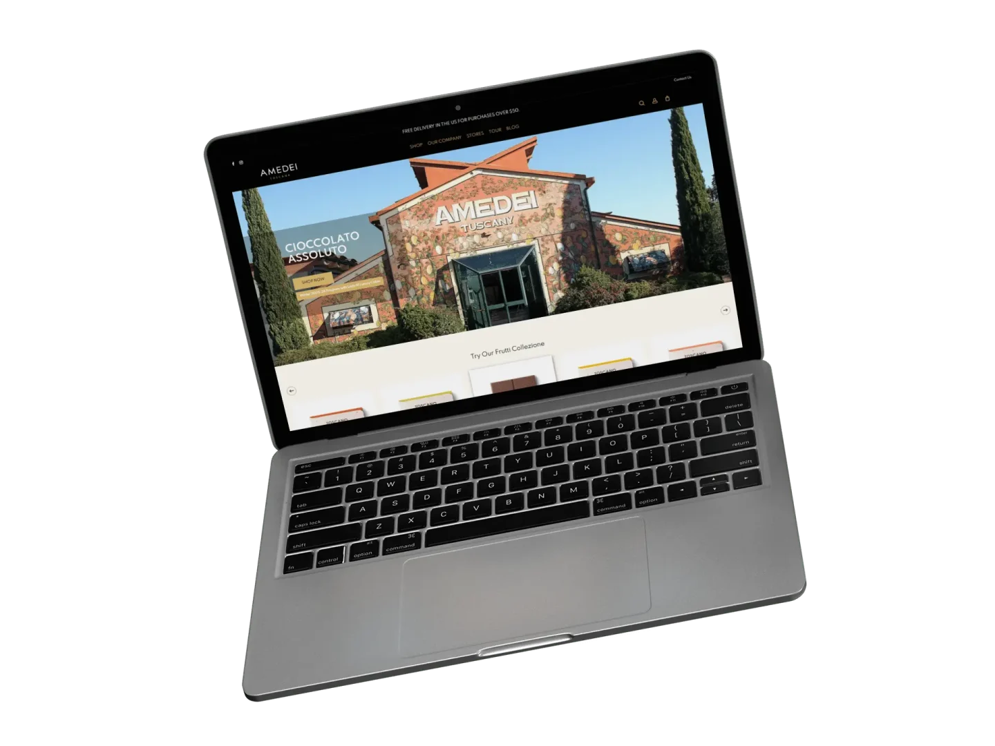 A silver laptop displays the Amedei website, featuring a large image of a stone building with AMEDEI on the front and a chocolate assortment sign to the left. The laptop is set against a white background.