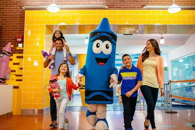 A family with three children walks indoors, smiling, alongside a person in a cheerful blue crayon costume. Bright yellow tiles and colorful decorations resembling crayons highlight the space’s playful spirit and brand purpose.