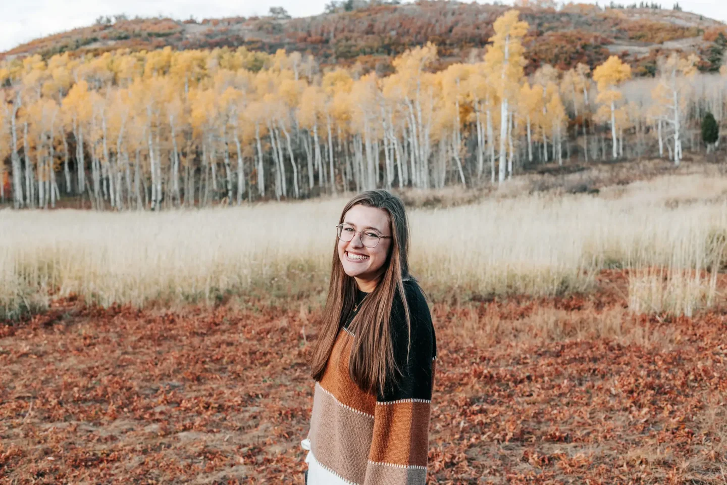 Natalie Nagy, a young woman with long brown hair and glasses, smiles while standing in a fall landscape with dry grass, autumn leaves, and trees with golden foliage in the background.