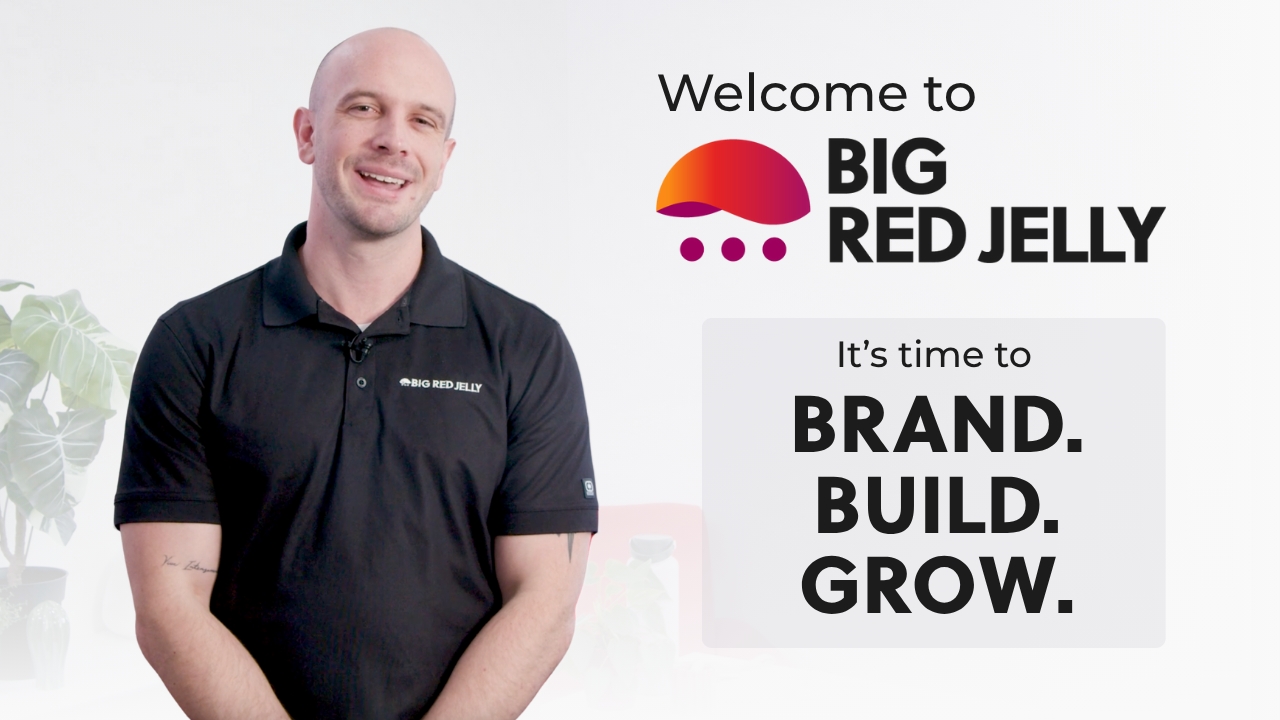 A smiling man in a black polo shirt stands next to a Welcome to Big Red Jelly sign with a red and orange logo. Text reads, It’s time to BRAND. BUILD. GROW. with potted plants in the background, highlighting a powerful digital marketing strategy.