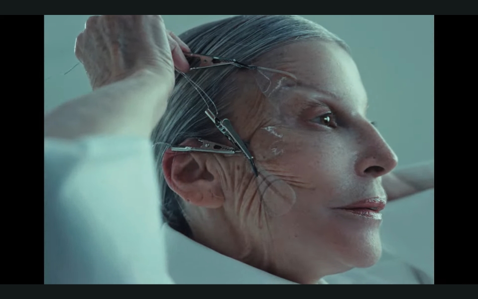 A person with gray hair pulls back their facial skin using metal clips and wires, revealing the contours and texture of their face. The scene has a clinical, surreal atmosphere.