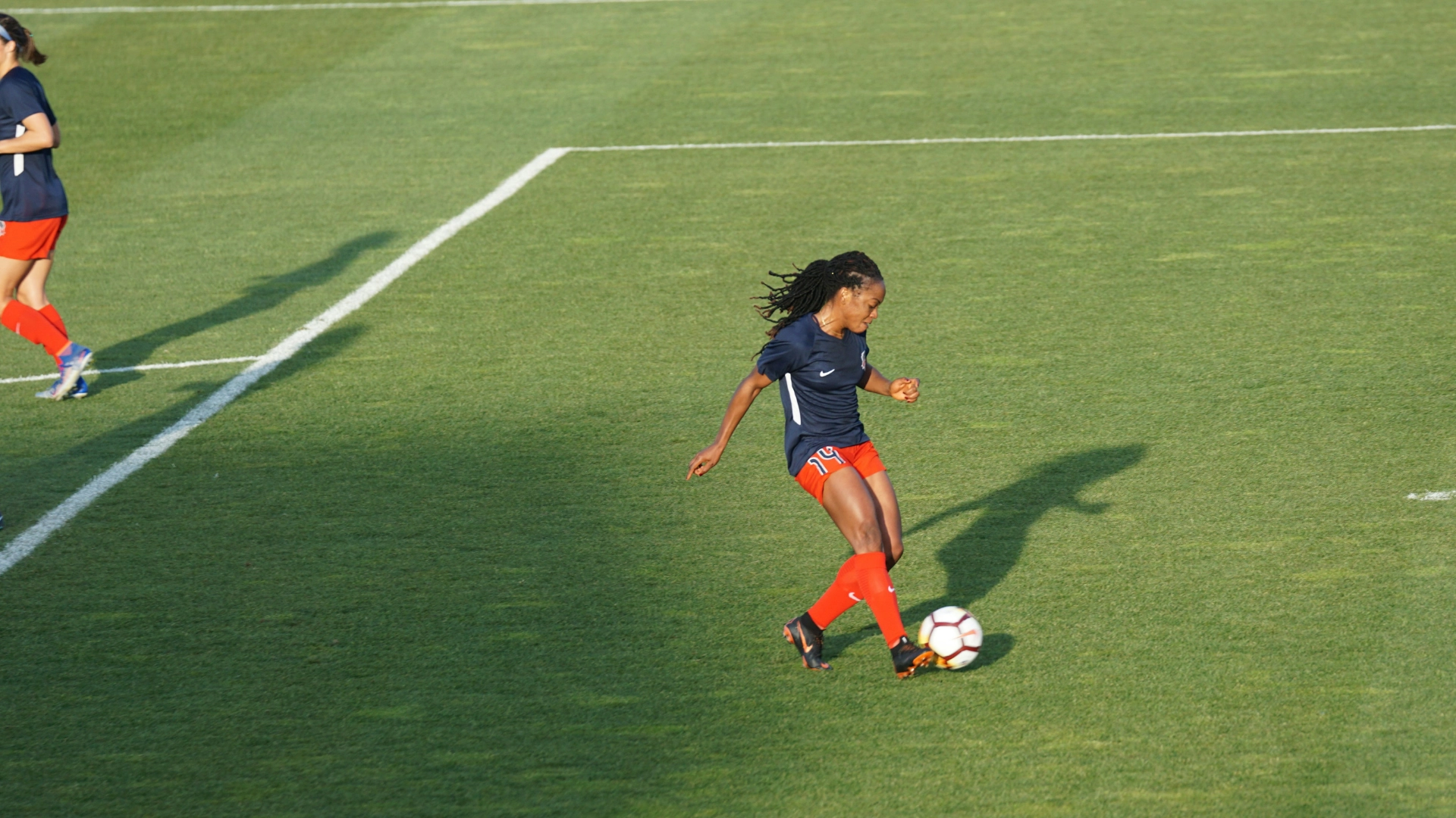 A soccer player in a navy shirt and red shorts prepares to kick a ball on a grassy field, while another player is partially visible in the background.