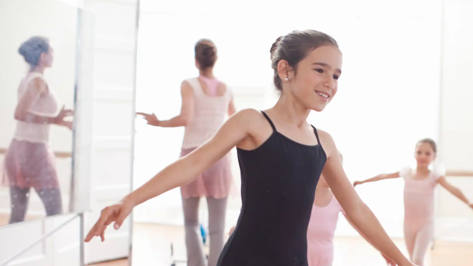 A young girl in a black leotard smiles while practicing ballet, arms outstretched. Other dancers and an instructor are visible in the bright, mirrored studio background.