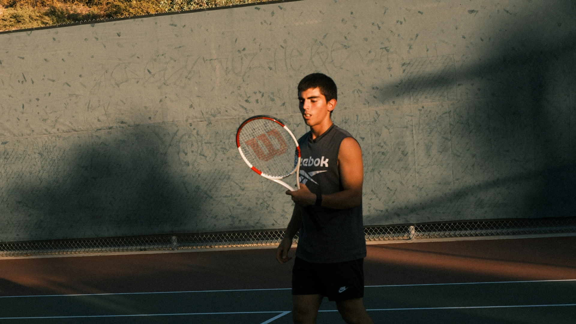 A young man in athletic clothing holds a tennis racket on an outdoor court, looking focused. The court is shaded, with a tall fence and greenery in the background.