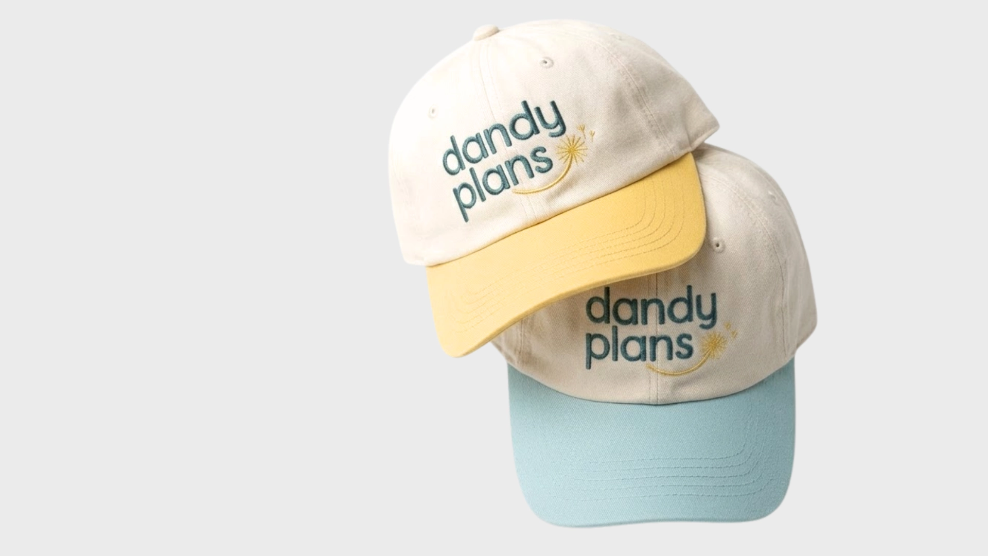 Two cream-colored baseball caps, both embroidered with dandy plans and a small yellow flower. One has a yellow brim, the other a light blue brim, and they are stacked on a plain, light gray background.