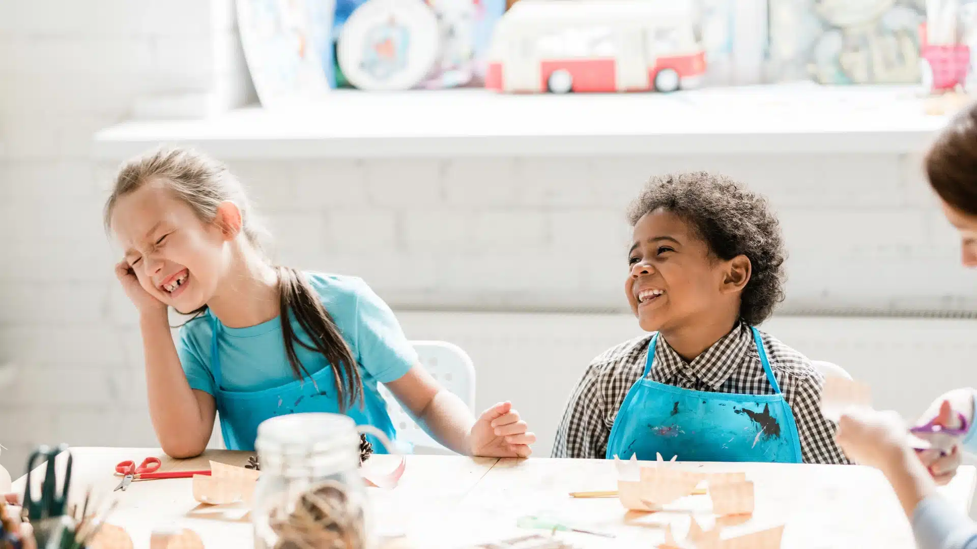 Two young children sit at a table, smiling and laughing together while wearing blue aprons, surrounded by craft supplies in a bright, cheerful room.