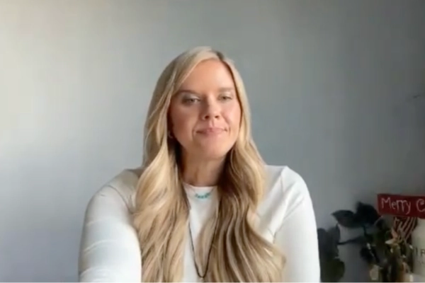 A woman with long blonde hair wearing a white top sits in front of a plain gray wall, with a plant and a small sign that reads Merry visible in the background.