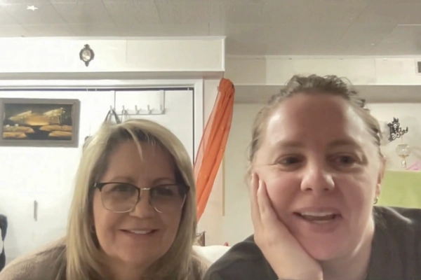 Two women are sitting indoors, smiling at the camera. One wears glasses and has blond hair, while the other has her hand on her cheek. The background shows a white wall, artwork, and an orange curtain.