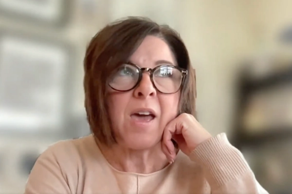 A woman with short brown hair and glasses sits indoors, resting her chin on her hand and looking slightly upward, wearing a light-colored top. The background is blurred.