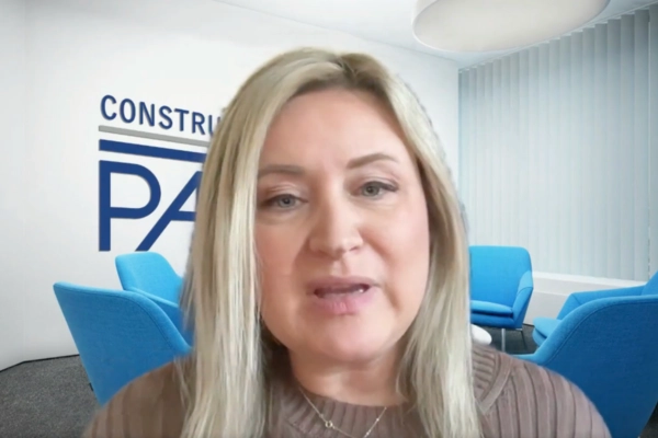 A woman with long blonde hair sits in a modern office with blue chairs and a logo on the wall reading CONSTRU PA. She is wearing a brown top and looking directly at the camera.