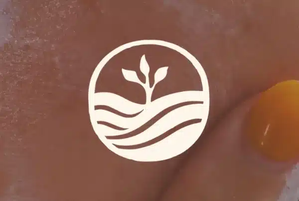 A hand with orange-painted nails applies cream to skin. Overlaid is the Primal Basics logo—a white circle featuring a sprouting plant above wavy lines.