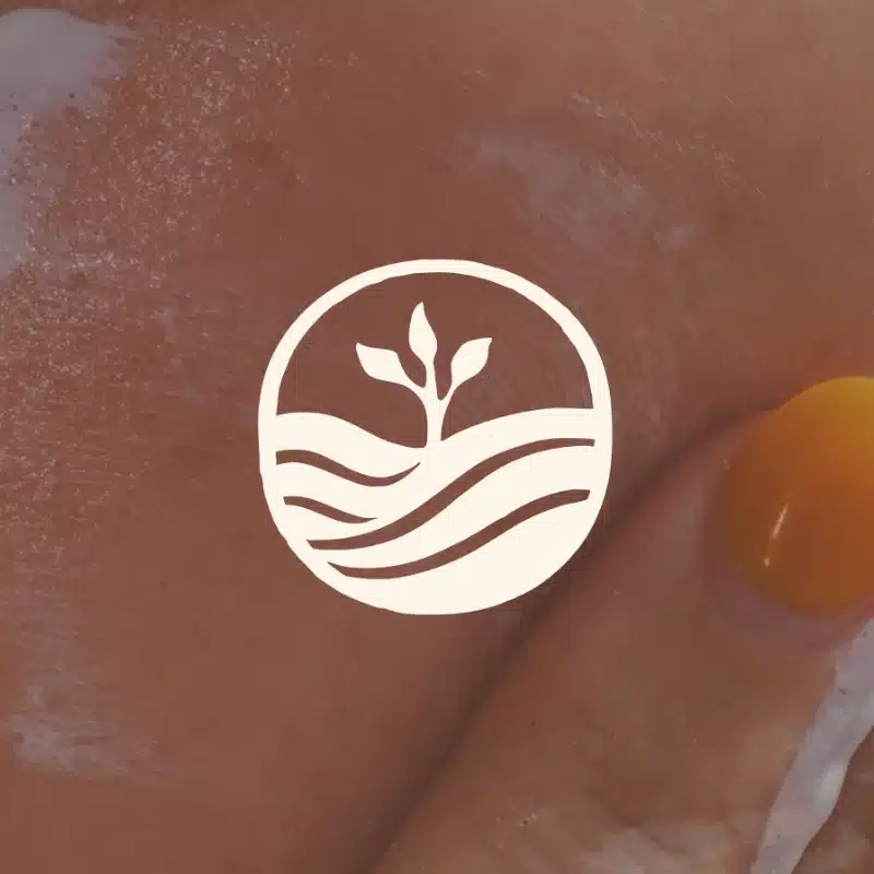 A hand with orange-painted nails applies cream to skin. Overlaid is the Primal Basics logo—a white circle featuring a sprouting plant above wavy lines.