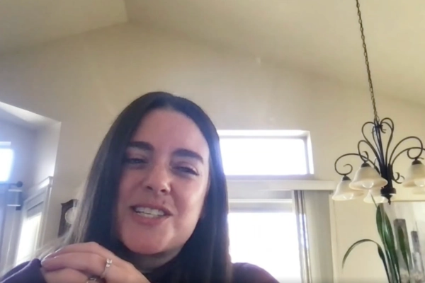 A woman with long brown hair smiles indoors, sitting in a bright room with large windows and a decorative hanging light fixture in the background.