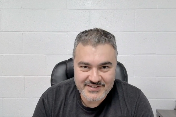 A man with short gray hair and a beard sits in a black chair, smiling at the camera. He is wearing a dark shirt and is positioned in front of a white brick wall.