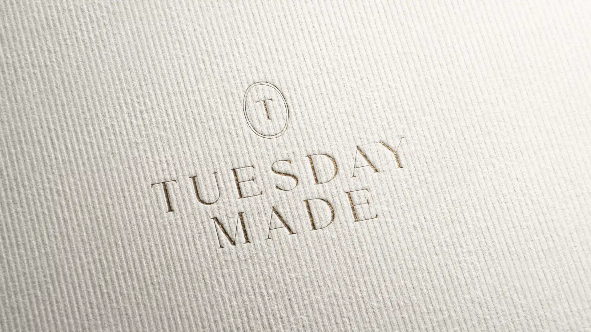 Embossed text on textured white paper reads TUESDAY MADE with a small circular emblem containing the letter T above the words.