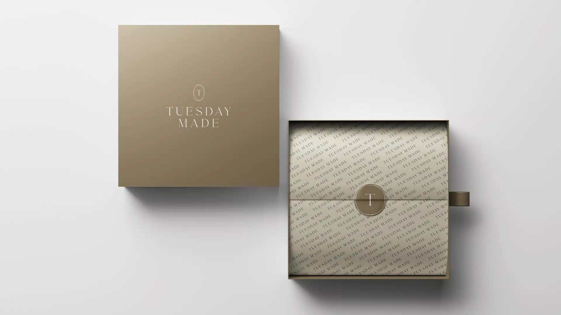 A square, beige gift box with TUESDAY MADE printed on the lid sits open, revealing neatly folded patterned paper inside, sealed with a circular sticker featuring a T logo. The box is on a white surface.