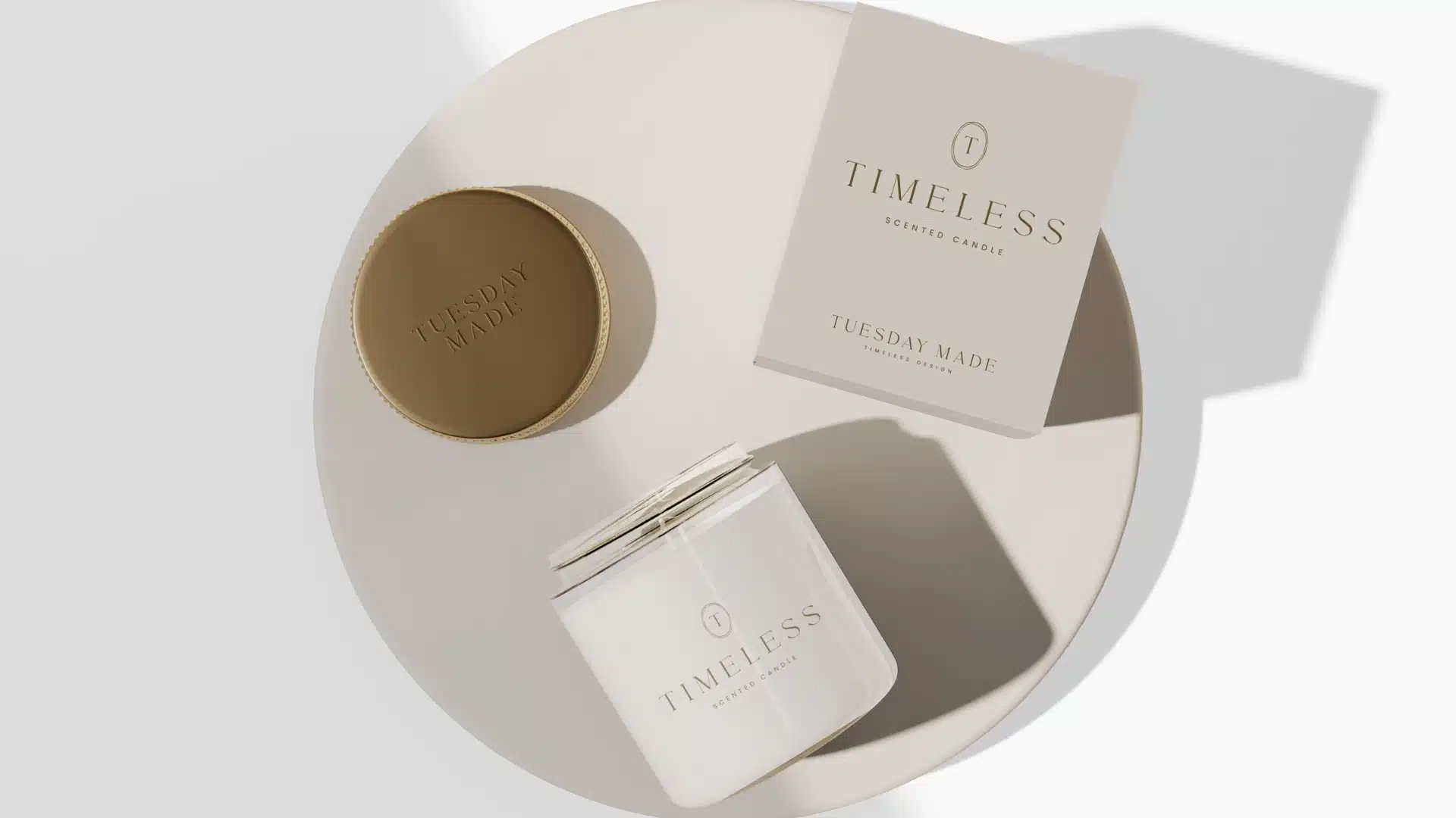 A white candle labeled TIMELESS with a gold lid and a matching box sit on a round, beige surface. Both the lid and the box feature the brand name TUESDAY MADE. The scene is brightly lit with soft shadows.