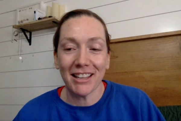 A woman with light skin and brown hair pulled back is smiling. She is wearing a blue sweatshirt and sitting in a room with white shiplap walls, a wooden headboard, and a shelf with various items behind her.