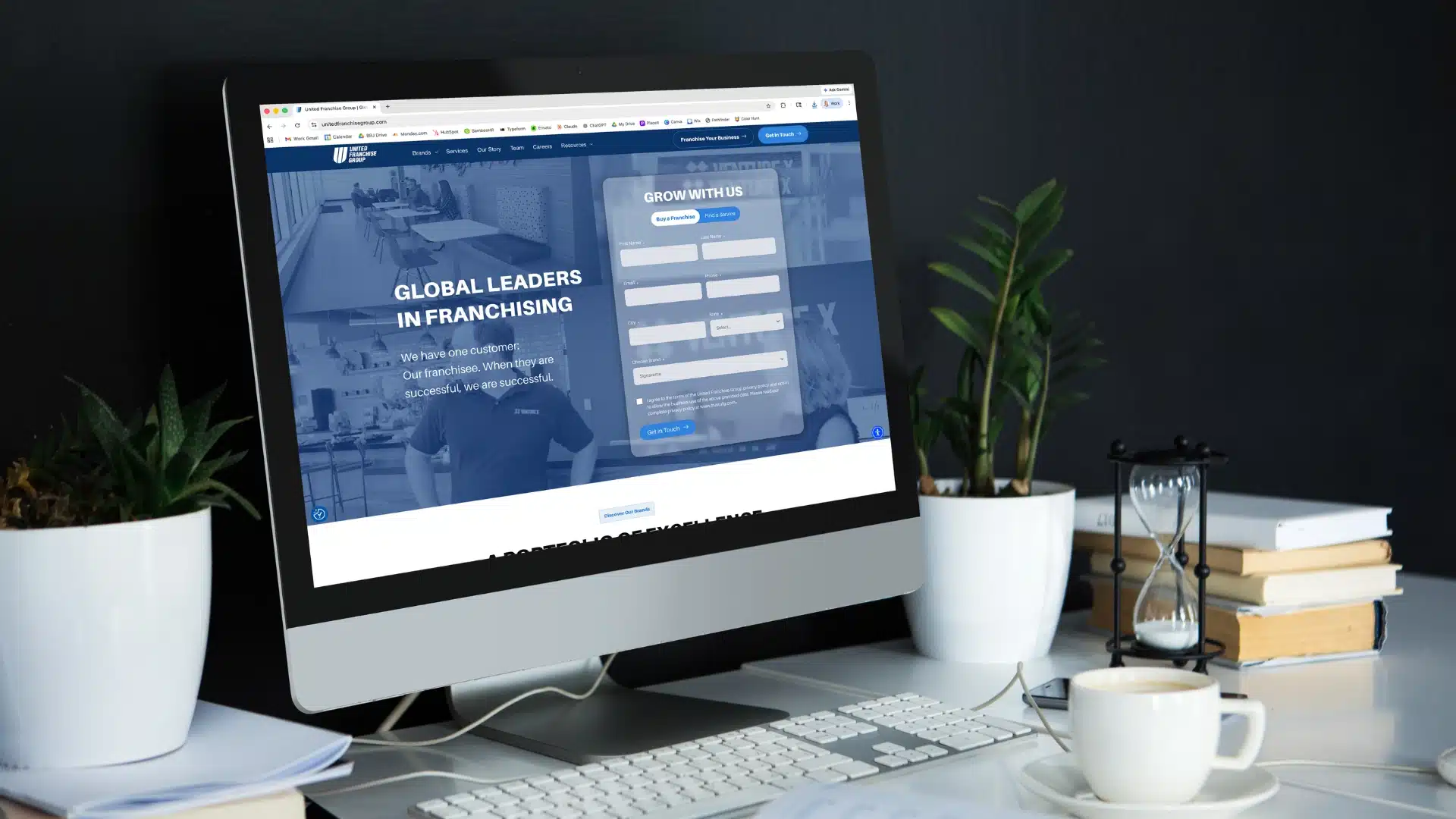 A desktop computer on a black desk displays a franchising website. The desk has a keyboard, mouse, white mug, potted plants, books, and an hourglass. The background is dark, creating a modern workspace atmosphere.