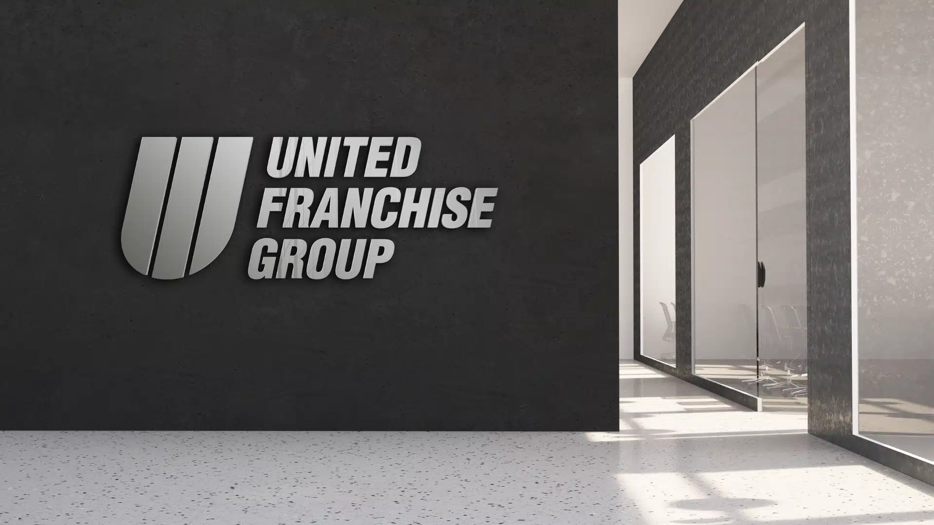 A modern office space with a dark wall featuring the silver logo and text United Franchise Group. Glass walls reveal a conference room with chairs in the background.