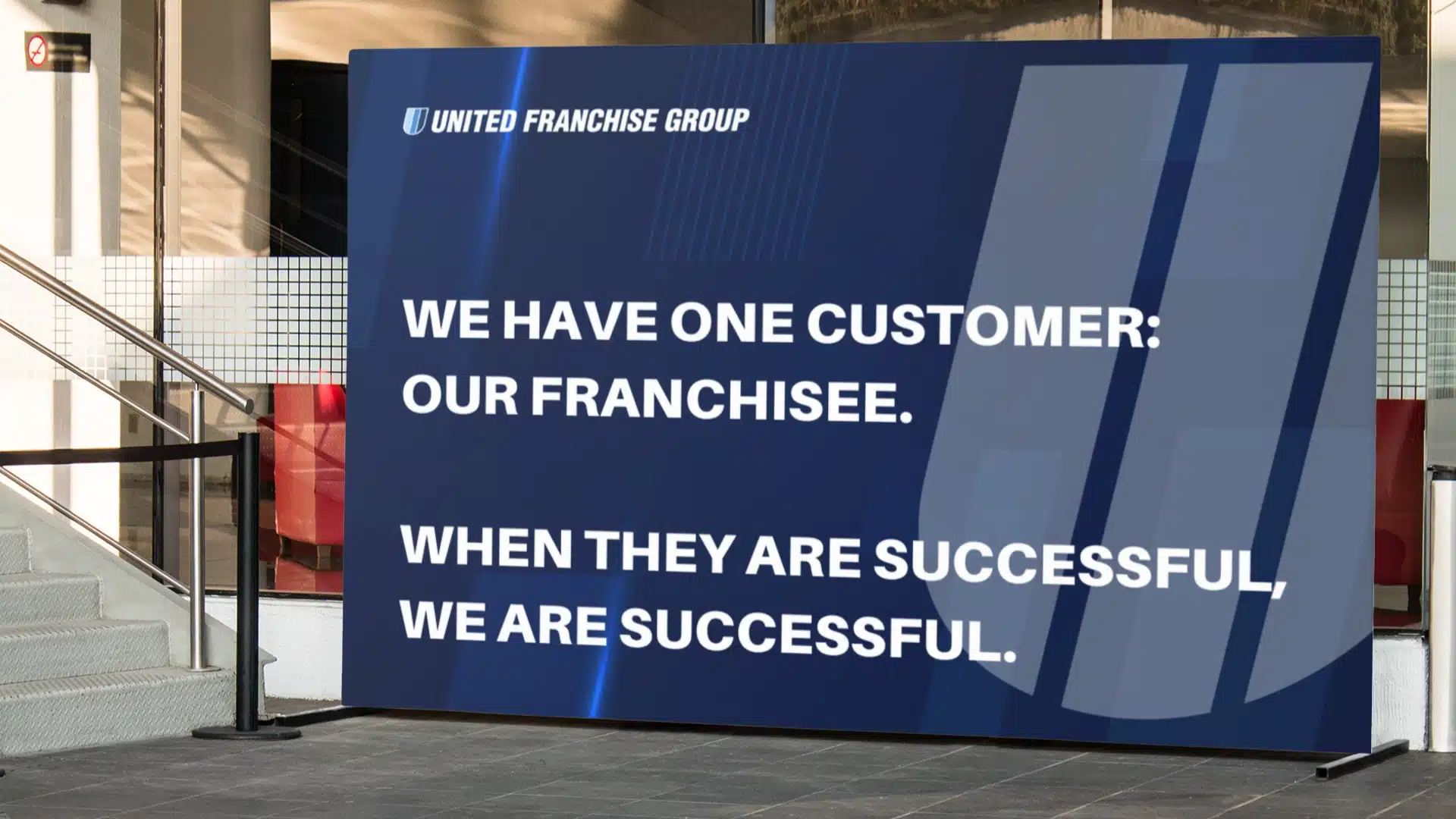 A large blue sign reads: “UNITED FRANCHISE GROUP. WE HAVE ONE CUSTOMER: OUR FRANCHISEE. WHEN THEY ARE SUCCESSFUL, WE ARE SUCCESSFUL.” The sign is displayed indoors beside a staircase and railing.