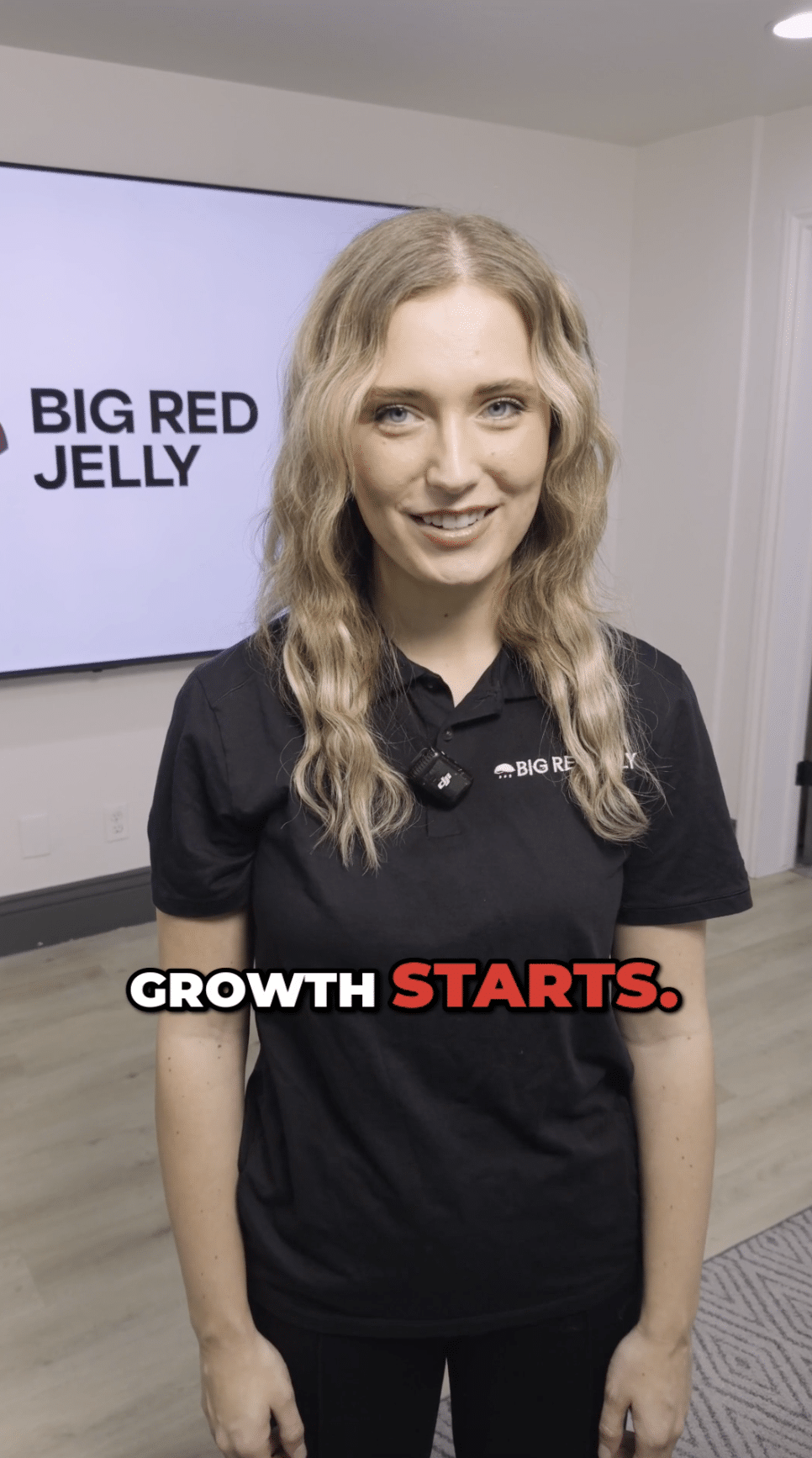 A woman with wavy blonde hair, wearing a black polo shirt, stands indoors smiling. Behind her is a screen displaying the Big Red Jelly logo. Text on the image reads: Growth starts.